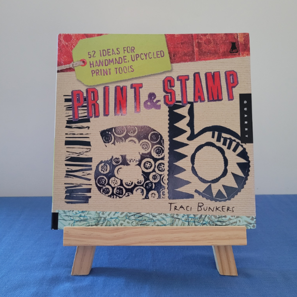 A book titled 'Print and Stamp' by Traci Bunkers displayed on a small wooden easel, with a colorful cover featuring various shapes and text.