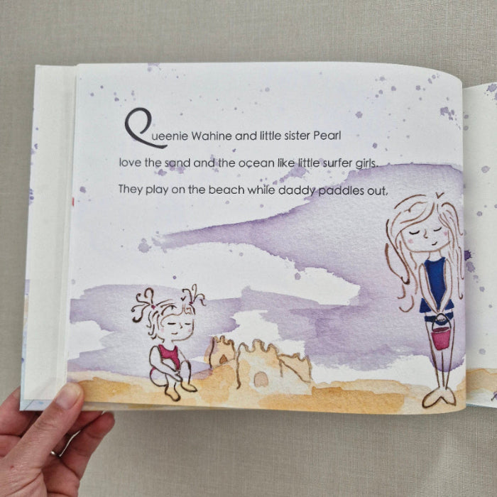 Illustrated book with characters playing on the beach, held by a hand against a neutral background.