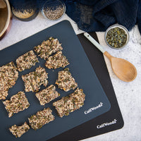 Reusable food-grade silicone baking mats with the brand name 'CaliWoods' printed on them, used for baking cookies, placed on a white surface with a bowl of oats and some nuts in the background.