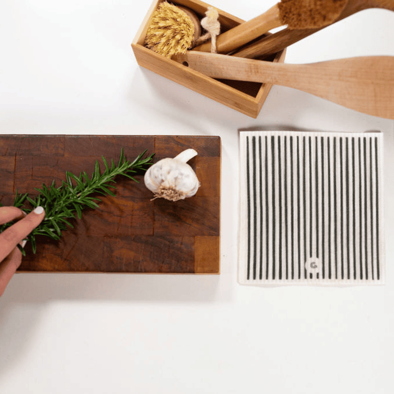 Wooden cutting board with herbs and garlic, wooden spoons, and a box of kitchen utensils on a white surface.