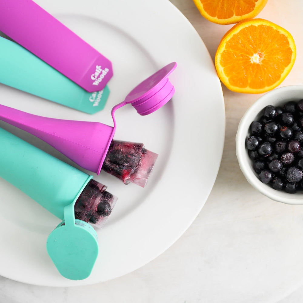 Colorful popsicle molds with popsicles on a plate, surrounded by oranges and blueberries.