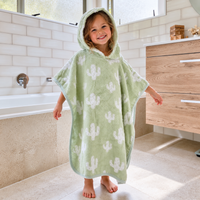 A young child wearing a light green hooded bath towel/ poncho with white cactus prints, standing in a bright bathroom.