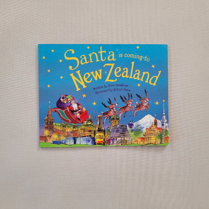A children's board book titled 'Santa is coming to New Zealand'  featuring a colorful illustrated cover with Santa in a red sleigh, kiwi birds, and a blue background, photographed on a light beige textured surface.