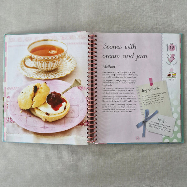 Cookbook open to a page with scones, cream, and jam illustration on a gray surface