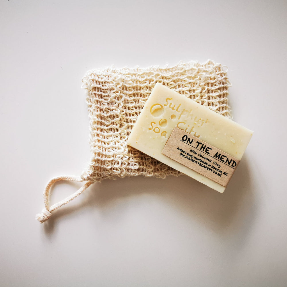 A beige soap saver bag made of sisal fibre, placed on a white surface with a soap 'On The Mend'.