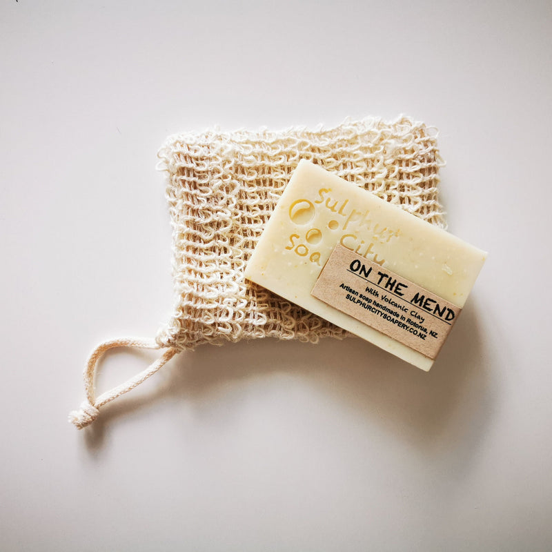 A beige soap saver bag made of sisal fibre, placed on a white surface with a soap 'On The Mend'.