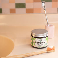 Solid toothpaste jar with a scoop and toothbrush on a bathroom counter.