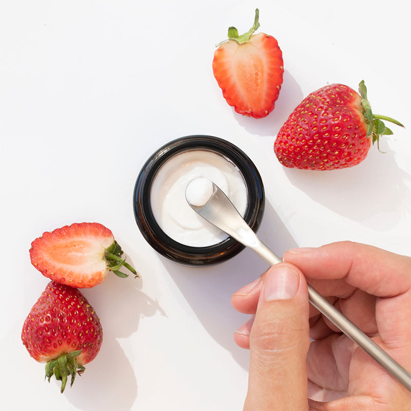 Hand using a scoop to get toothpaste from a jar with strawberries on a white background