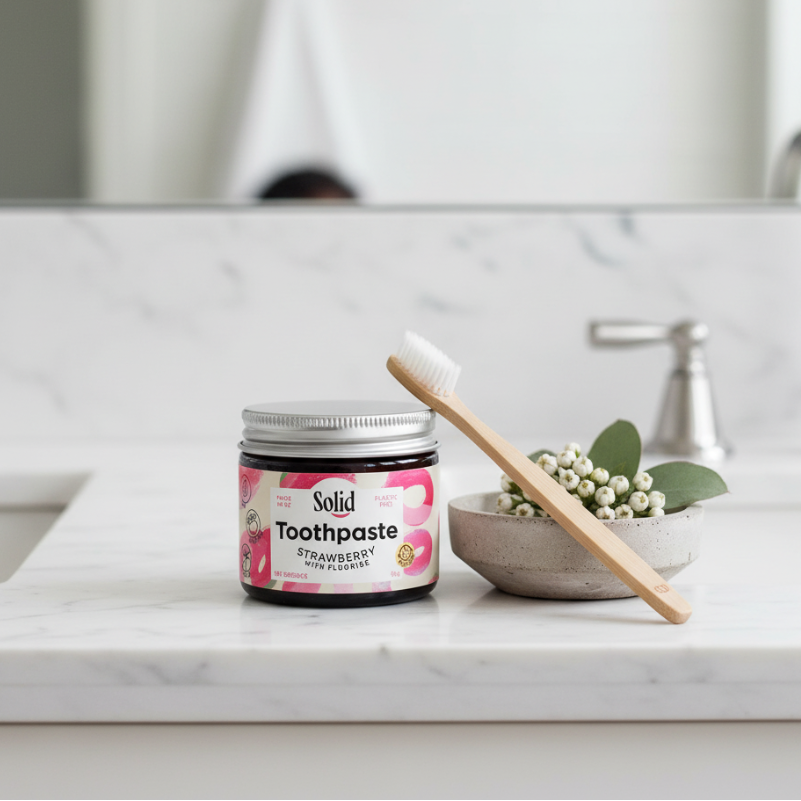 Jar of strawberry-flavoured solid toothpaste with a visible brand label sitting on a bathroom counter with wooden toothbrush