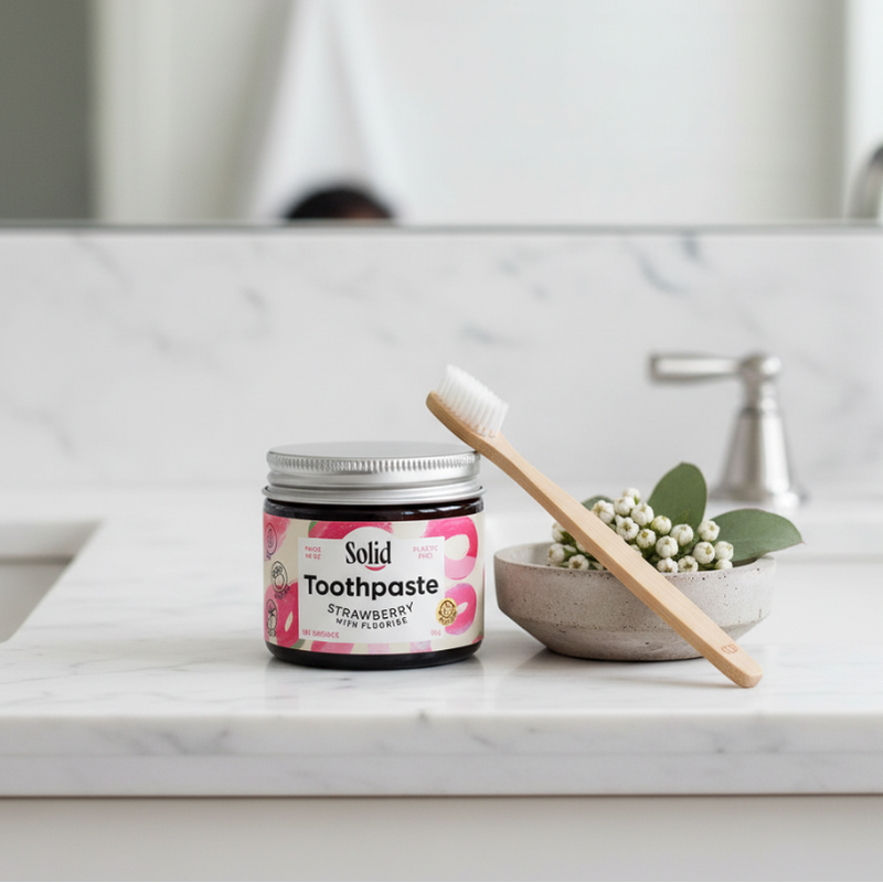 Jar of strawberry-flavoured solid toothpaste with a visible brand label sitting on a bathroom counter with wooden toothbrush