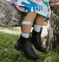 Child wearing black boots with a bandage on their knee, sitting against a tree.