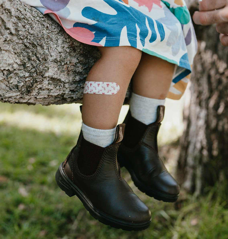 Child wearing black boots with a bandage on their knee, sitting against a tree.
