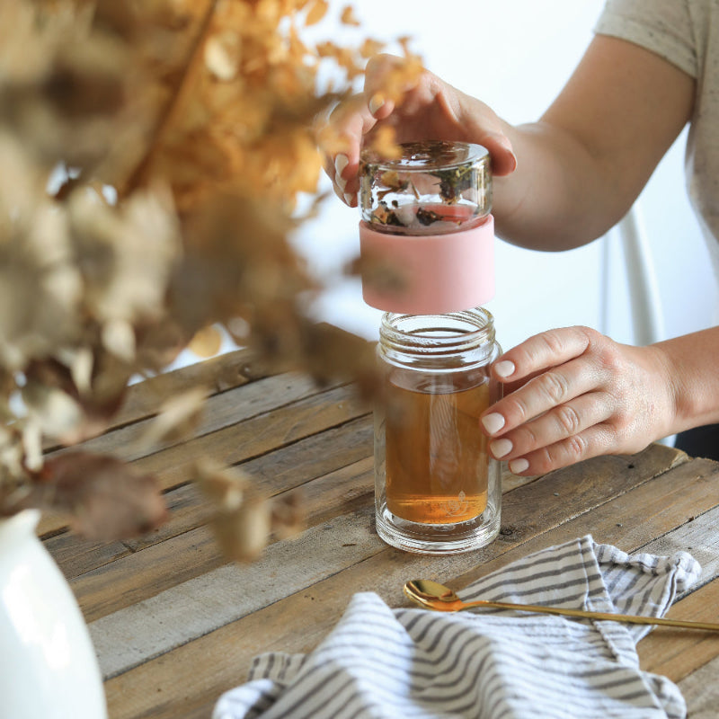 Person holding a glass flask of tea with a pink sleeve on a wooden table