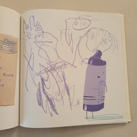 An open sketchbook or art book showing a childlike purple drawing of a bottle and scribbled figures on cream pages, with a torn beige paper fragment visible at the bottom.
