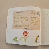 An open book or notebook showing a vintage-style postcard page with illustrated drawings and handwritten text on cream paper, photographed on a beige fabric surface.