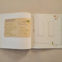 An open book or journal laid flat on a beige fabric surface, showing illustrated pages with a tan postcard-like drawing on the lower page and simple line drawings on the upper page.