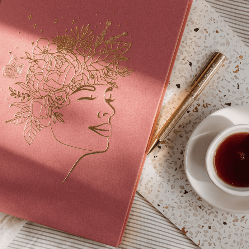 Blush Journal with gold embossed design of a face and flowers, next to a gold luxe pen and cup of tea on a textured surface.