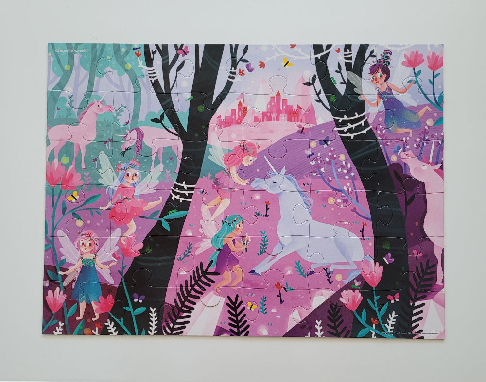A completed children's jigsaw puzzle featuring a whimsical fantasy forest scene with fairies, unicorns, trees, flowers, and a pink castle, photographed against a light neutral background.