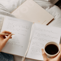 Person writing in 'YOU' wellbeing journal with a cup of coffee on a textured surface
