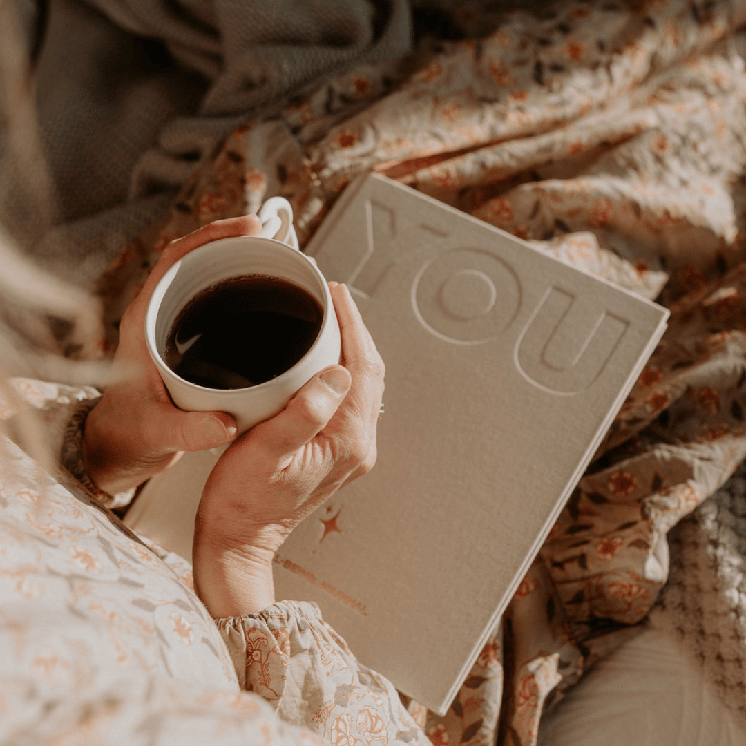 Person holding a cup of coffee next to a book titled 'YOU' on a floral blanket.