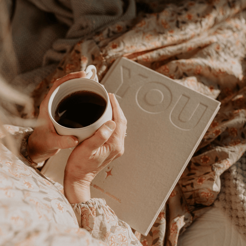 Person holding a cup of coffee next to a book titled 'YOU' on a floral blanket.