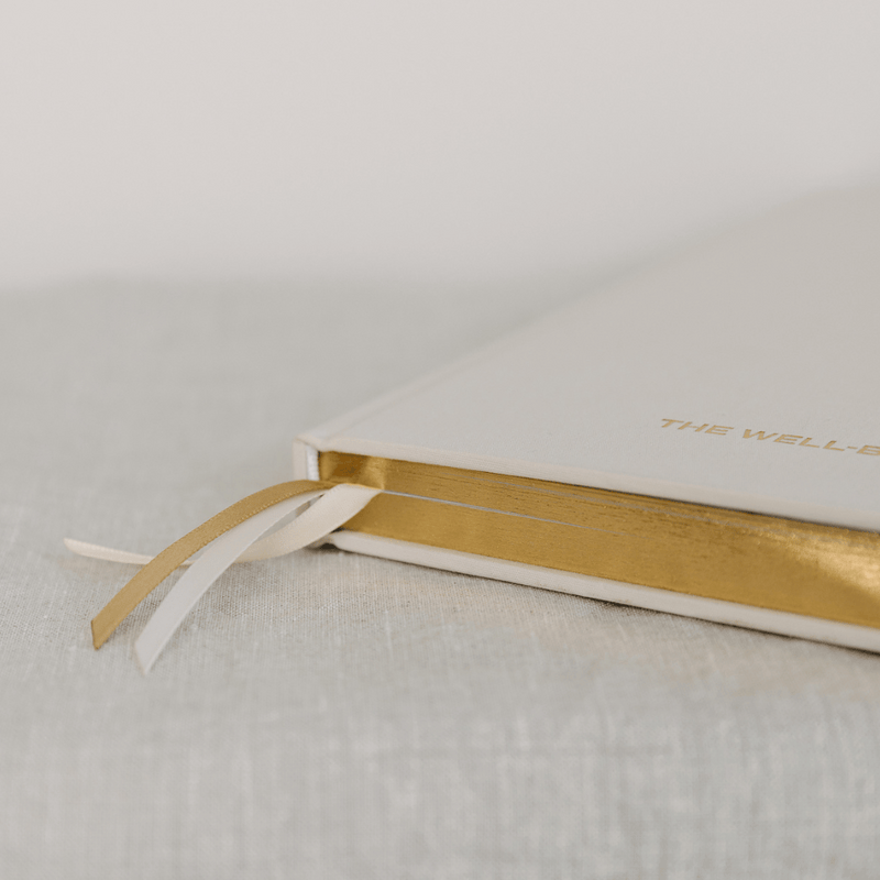 Linen covered book in sand with gold spine and triple ribbons on a textured surface