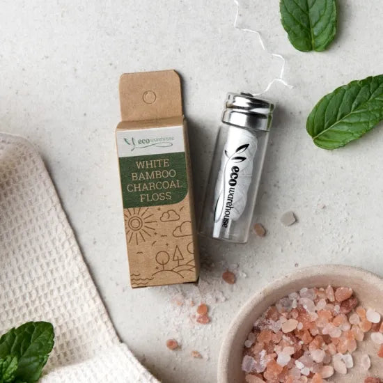 A small bamboo charcoal floss bottle with its kraft paper box, styled on a light textured surface with mint leaves, a white cloth, and a bowl of pink Himalayan salt. The box reads 'eco environment' and 'WHITE BAMBOO CHARCOAL FLOSS'.