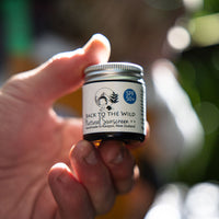 Hand holding a small jar of Back to the Wild natural sunscreen with a blurred outdoor background