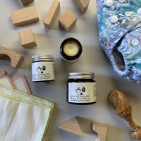 The image shows a small open jar of 'Nappy Barrier Balm' with its contents partially visible, placed next to a wooden block and a cloth with a pattern. In the background, there's a diaper and a wooden spoon lying on the surface.