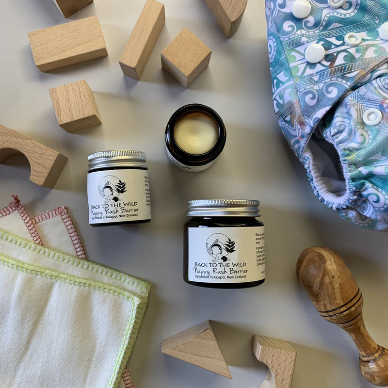 The image shows a small open jar of 'Nappy Barrier Balm' with its contents partially visible, placed next to a wooden block and a cloth with a pattern. In the background, there's a diaper and a wooden spoon lying on the surface.