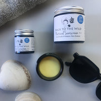 A glass jar of natural sunscreen labeled 'Back to the Wild Natural Sunscreen SPF 50+' alongside a smaller jar, a towel, a shell, and a pair of sunglasses on a light background.